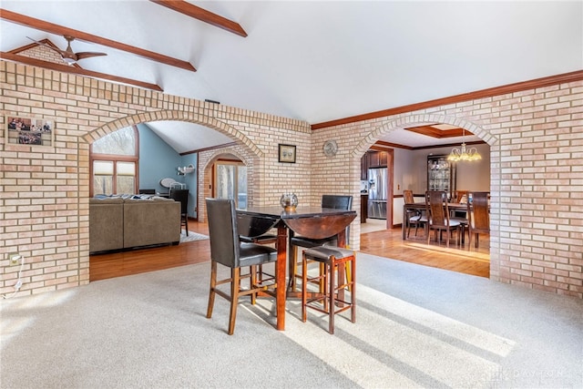 carpeted dining space with ceiling fan with notable chandelier, lofted ceiling with beams, and brick wall