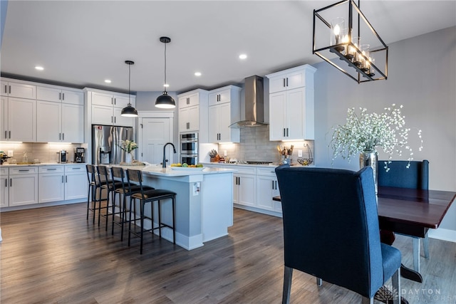 kitchen featuring dark hardwood / wood-style flooring, an island with sink, pendant lighting, wall chimney range hood, and white cabinets