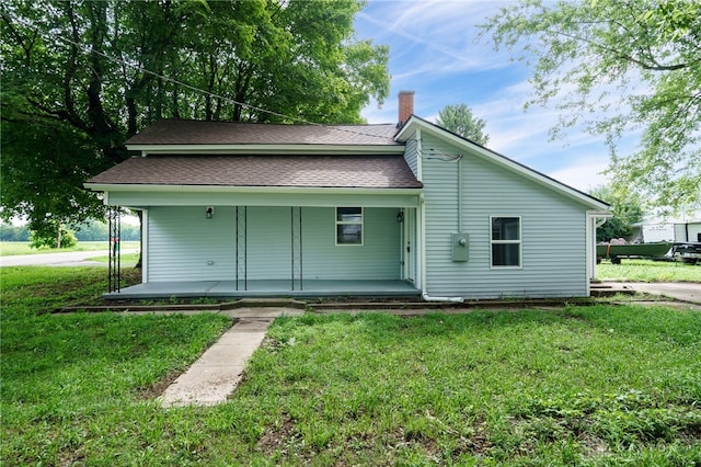 back of house featuring a lawn, covered porch, a chimney, and roof with shingles