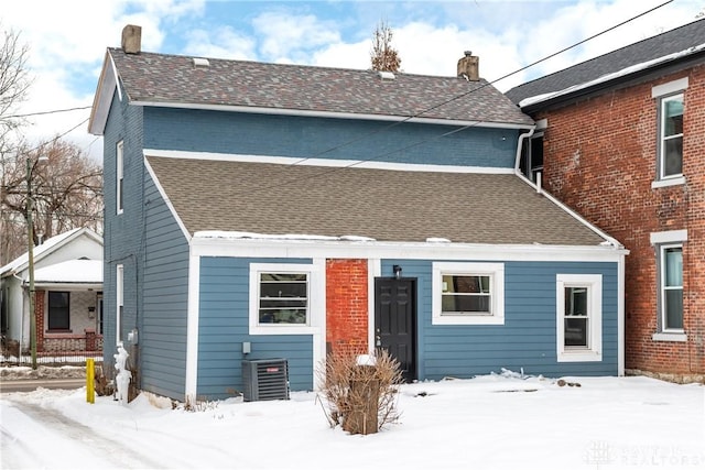 snow covered property featuring central air condition unit, roof with shingles, and a chimney