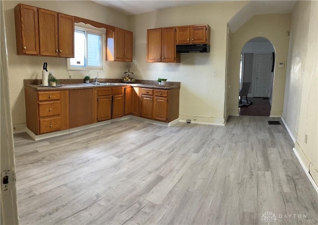 kitchen with arched walkways, dark countertops, brown cabinetry, light wood-style floors, and under cabinet range hood