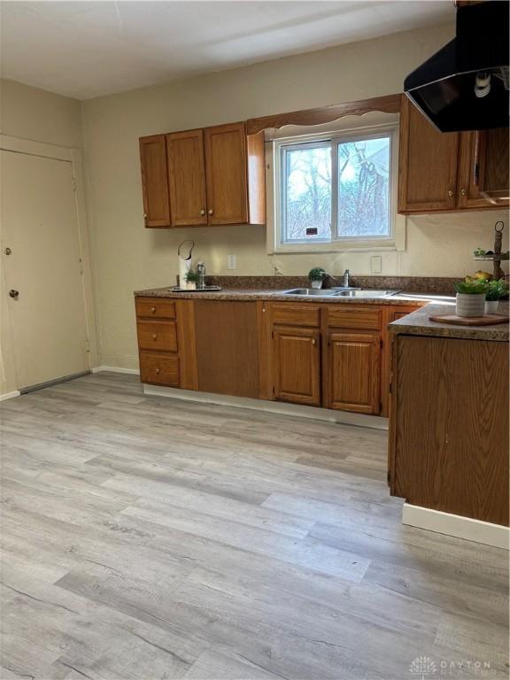 kitchen with light wood finished floors, brown cabinetry, dark countertops, a sink, and exhaust hood