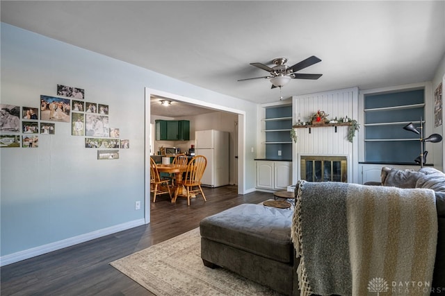 living area featuring dark wood-style floors, built in features, a fireplace, ceiling fan, and baseboards