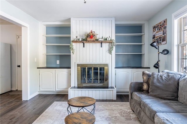 living room with dark wood-type flooring, a glass covered fireplace, baseboards, and built in shelves