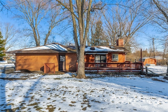 snow covered back of property featuring brick siding, a chimney, a storage shed, a deck, and an outdoor structure