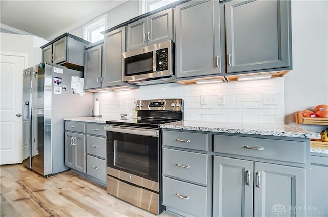 kitchen with gray cabinetry, stainless steel appliances, light stone countertops, decorative backsplash, and light wood-type flooring