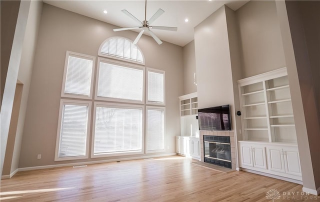 unfurnished living room with a high ceiling, a tiled fireplace, ceiling fan, and light hardwood / wood-style flooring