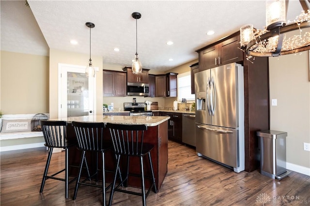 kitchen featuring dark brown cabinetry, decorative light fixtures, a kitchen island, stainless steel appliances, and light stone countertops