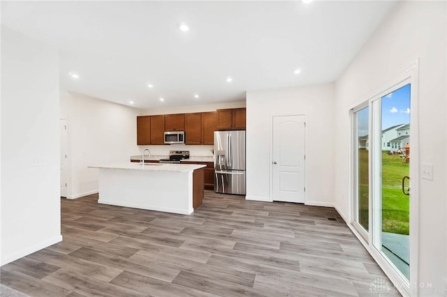 kitchen with an island with sink, sink, stainless steel appliances, light stone countertops, and light wood-type flooring