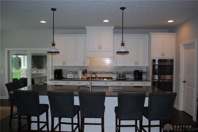 kitchen with decorative light fixtures, double oven, an island with sink, decorative backsplash, and white cabinets