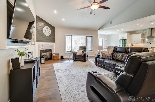 living room with sink, high vaulted ceiling, light wood-type flooring, a tile fireplace, and ceiling fan