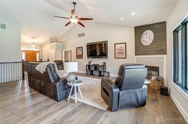 living room with ceiling fan with notable chandelier, high vaulted ceiling, a tiled fireplace, light hardwood / wood-style floors, and plenty of natural light