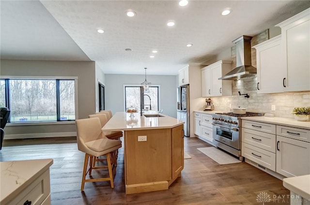 kitchen with wall chimney exhaust hood, sink, appliances with stainless steel finishes, a kitchen island with sink, and white cabinets