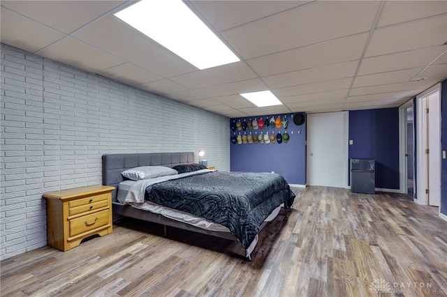 bedroom with a drop ceiling, hardwood / wood-style flooring, refrigerator, and brick wall