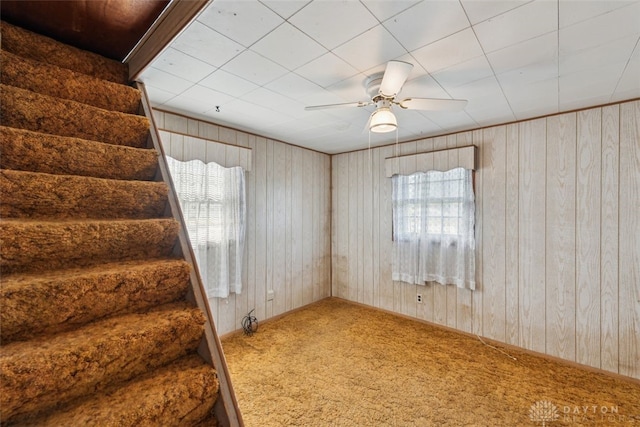 carpeted empty room featuring wood walls, ceiling fan, stairs, and a wealth of natural light