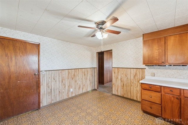 kitchen with a ceiling fan, light countertops, wainscoting, brown cabinetry, and wallpapered walls