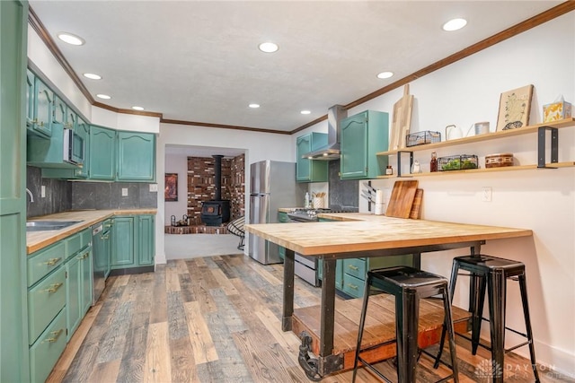 kitchen featuring sink, appliances with stainless steel finishes, butcher block counters, wall chimney exhaust hood, and a wood stove