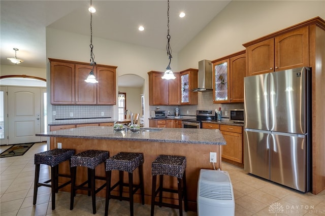 kitchen with hanging light fixtures, an island with sink, appliances with stainless steel finishes, and wall chimney exhaust hood