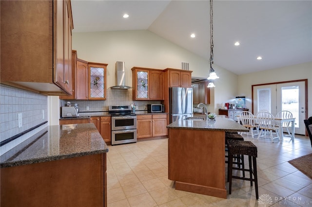 kitchen with an island with sink, sink, hanging light fixtures, stainless steel appliances, and wall chimney exhaust hood