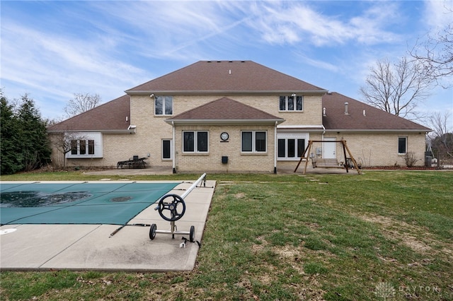 rear view of property with a covered pool, roof with shingles, brick siding, a patio, and a yard