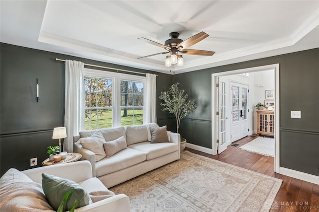 living area with crown molding, ceiling fan, a raised ceiling, wood-type flooring, and baseboards