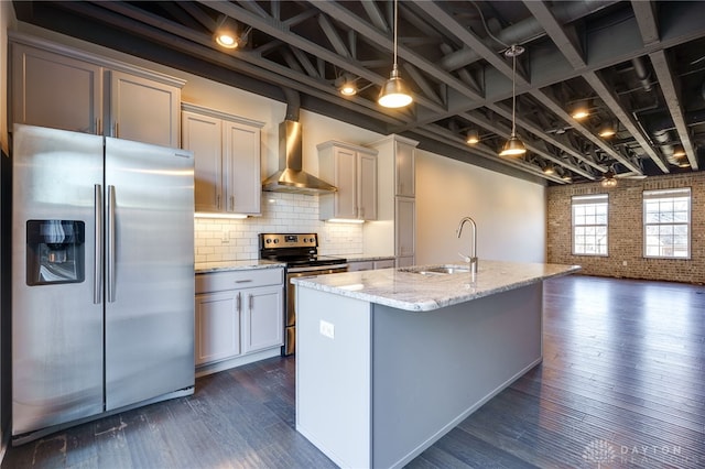 kitchen with wall chimney range hood, appliances with stainless steel finishes, brick wall, light stone counters, and a center island with sink