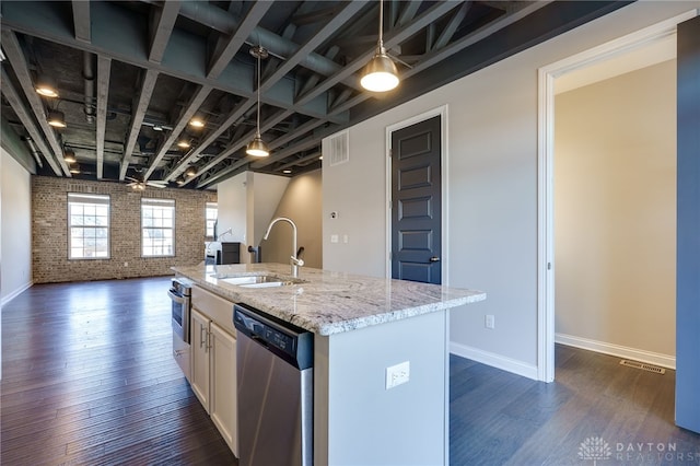 kitchen with brick wall, appliances with stainless steel finishes, an island with sink, sink, and light stone countertops