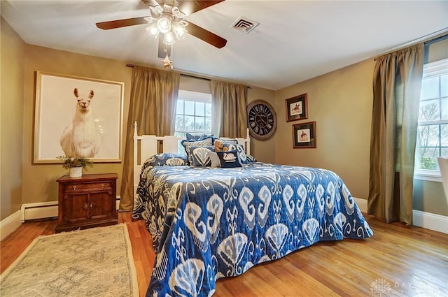 bedroom with multiple windows, baseboard heating, wood-type flooring, and ceiling fan