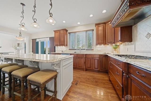 kitchen with light stone counters, brown cabinets, stainless steel gas cooktop, decorative light fixtures, and custom range hood