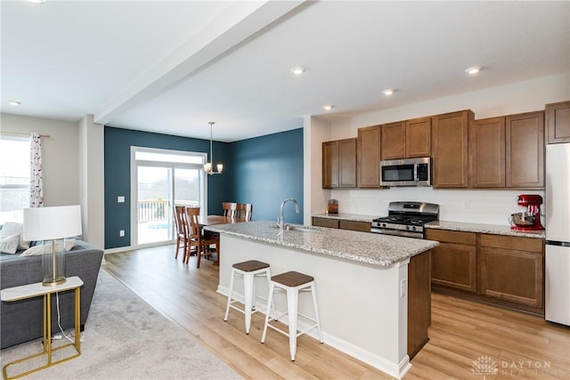 kitchen with decorative light fixtures, appliances with stainless steel finishes, brown cabinetry, a kitchen island with sink, and a sink