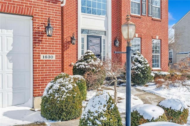 snow covered property entrance with an attached garage and brick siding