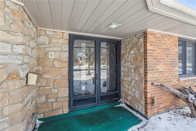 snow covered property entrance featuring stone siding, french doors, and brick siding