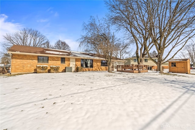 snow covered rear of property with a deck, an outbuilding, central AC, brick siding, and a shed