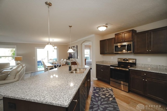 kitchen with arched walkways, a sink, dark brown cabinets, appliances with stainless steel finishes, and a center island with sink