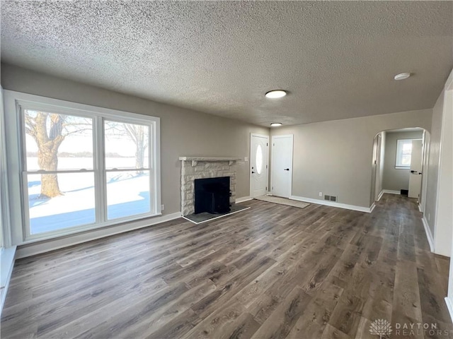 unfurnished living room with arched walkways, a stone fireplace, visible vents, baseboards, and dark wood-style floors