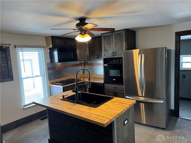 kitchen with a kitchen island with sink, dark brown cabinetry, a sink, butcher block countertops, and black appliances