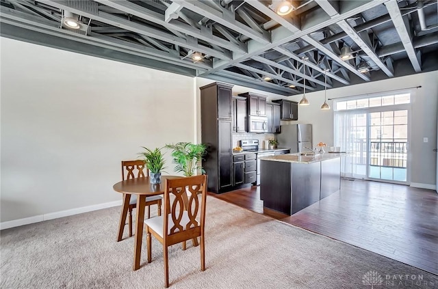 kitchen with light stone counters, appliances with stainless steel finishes, a kitchen island with sink, dark brown cabinets, and baseboards