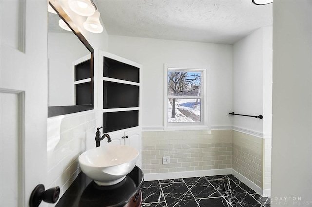 bathroom featuring marble finish floor, a wainscoted wall, tile walls, a sink, and a textured ceiling