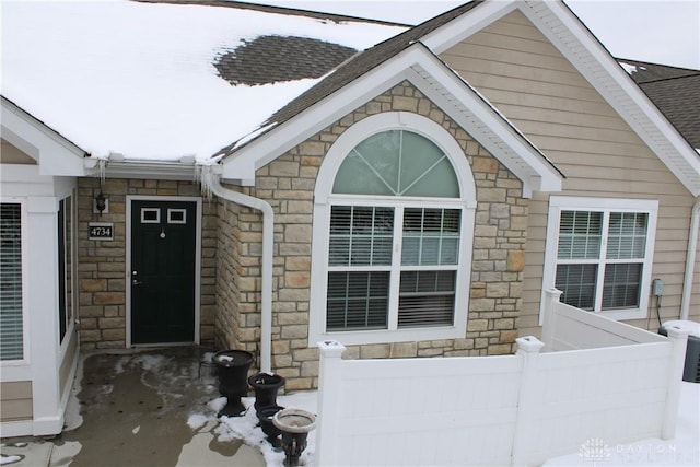 snow covered property entrance with a shingled roof, stone siding, and fence