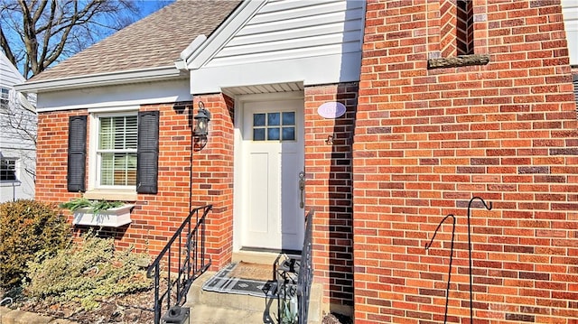 doorway to property featuring brick siding and roof with shingles