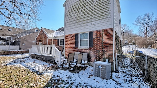 snow covered property featuring central air condition unit, a wooden deck, fence, and brick siding