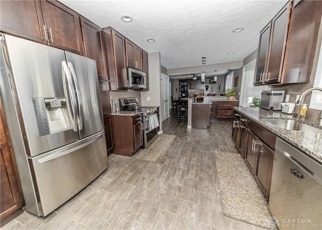 kitchen with dark brown cabinetry, appliances with stainless steel finishes, light stone countertops, a textured ceiling, and a sink