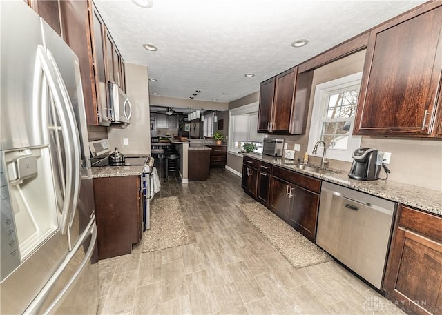 kitchen featuring light stone counters, stainless steel appliances, a sink, a textured ceiling, and dark brown cabinets