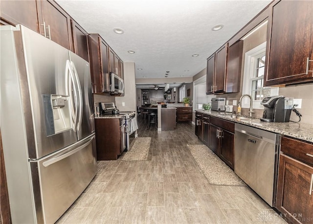 kitchen featuring light stone counters, recessed lighting, appliances with stainless steel finishes, dark brown cabinetry, and a sink