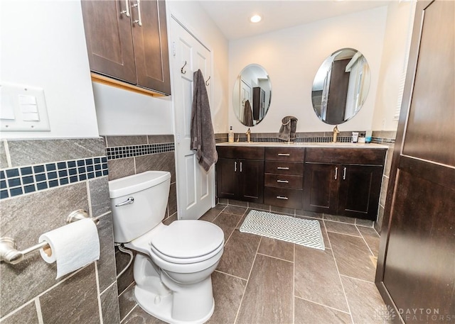 bathroom with toilet, recessed lighting, a wainscoted wall, tile walls, and double vanity