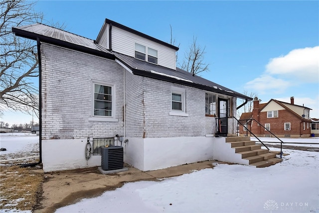 snow covered property featuring metal roof, central AC, and brick siding