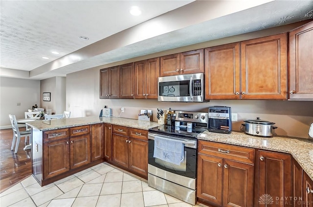 kitchen featuring stainless steel appliances, brown cabinetry, a peninsula, and light stone countertops