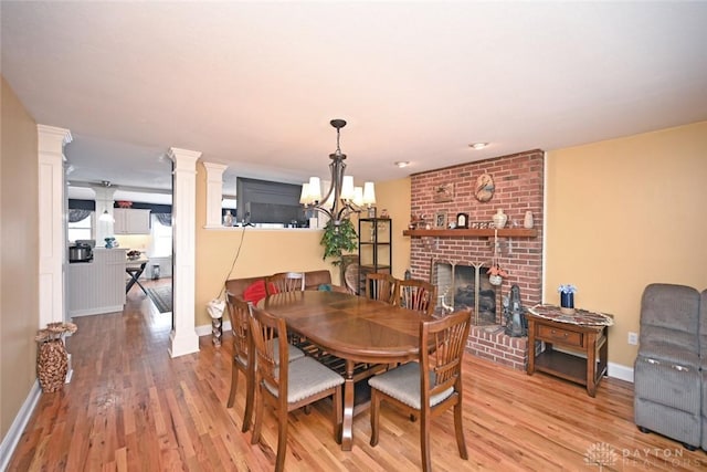 dining area with a notable chandelier, baseboards, light wood-type flooring, a brick fireplace, and decorative columns