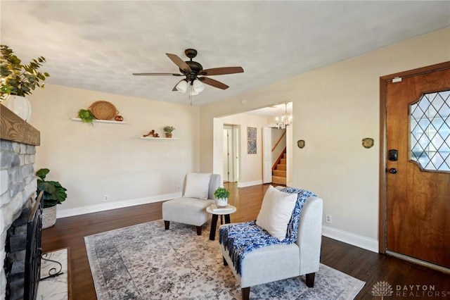 sitting room with dark wood-style flooring, stairway, a ceiling fan, a stone fireplace, and baseboards