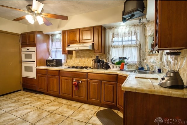 kitchen featuring tasteful backsplash, white double oven, under cabinet range hood, stainless steel gas stovetop, and a sink